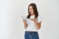 Young woman looking angry and confused at smartphone screen, complaining on broken app, standing over white background