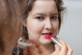 Young woman is looking in mirror and applying red glossy lipstick on her lips