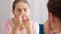 Young woman looking in mirror and squeezing pimples on her nose. Concept of female aging, skin issues, facial treatment and care Royalty Free Stock Photo