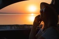 Young woman looking out car window of the sunset on the sea and talking on the phone. Royalty Free Stock Photo