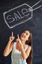 Young woman looking up and pointing at big chalk sale inscription on blackboard