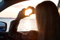 A young woman looks out the car window at the sunset on the sea.heart made with hands