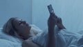 Young woman lying in the bed with a smartphone at night. Beautiful blond resting girl. Twilight in the bedroom