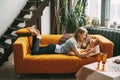 A young woman is lying on the sofa and reading a favorite book. The girl is resting, devotes her free time to reading. Side view Royalty Free Stock Photo