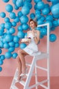 A young woman makes a wish with cake sitting on stepladder on pink wall background with blue bubbles. 20s candles Royalty Free Stock Photo