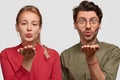 Young woman and man fold lips, hold palms near mouthes, blow air kiss at camera, have romantic look, love expression