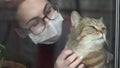 A young woman in a mask strokes a cat. The girl in quarantine at home stands at the window and caresses the British cat