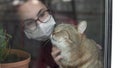 A young woman in a mask strokes a cat. The girl in quarantine at home stands at the window and caresses the British cat