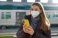 Young woman with medical face mask buying ticket online with smartphone on train station Royalty Free Stock Photo
