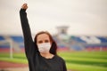 Young woman in medical mask doing warm-up at stadium. Adult female in black casual clothes doing sports in fresh air and