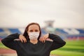 Young woman in medical mask doing warm-up at stadium. Adult female in black casual clothes doing sports in fresh air and