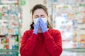 A young woman in a medical mask and gloves sneezes, covering her face. In the background, the shop Windows of pharmacy products.