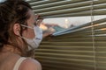 Young woman in medical mask looks out the window through the blinds Royalty Free Stock Photo