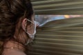 Young woman in medical mask looks out the window through the blinds Royalty Free Stock Photo