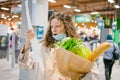 Young woman in a medical mask looks shocked at a paper check in a grocery supermarket holding a paper bag with groceries