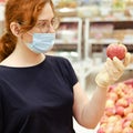 A young woman in a medical mask between rows of fruit in a supermarket. A girl buys apples in a store. Visiting public places Royalty Free Stock Photo