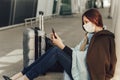 Young woman in medical mask sits near luggage in airport. Woman waiting for her flight, using a mobile phone Royalty Free Stock Photo