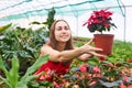 Young woman in the middle of a greenhouse smiling holds out a red flower in front of herself