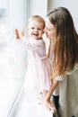 Young woman mom with long hair in cozy knitted cardigan with baby girl on hands near window at the home Royalty Free Stock Photo