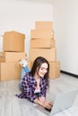Young woman is moving using a laptop and smiling, lying among cardboard boxes in new home Royalty Free Stock Photo