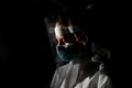 Young woman nurse in medical mask and protective screen looking down in dark room.