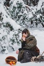 A young woman in old clothes is sitting with brushwood and a basket with apples in the snow in the winter forest Royalty Free Stock Photo