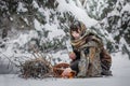 A young woman in old clothes is sitting with brushwood and a basket with apples in the snow in the winter forest