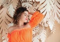 young woman in orane top posing on beige background with tropical paper leaves.