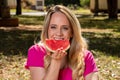 Young woman outdoors under trees in a park, happy and smiling, doing picnic and tasting watermelon and offering a slice