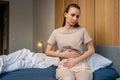 A young woman with a painful facial expression, holding her stomach, suffering from menstrual pains, is sitting on a Royalty Free Stock Photo