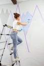 Full length view of young woman painting wall with paint roller and using masking tape while standing on ladder Royalty Free Stock Photo