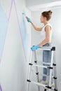 Young woman painting wall with paint roller and using masking tape while standing on ladder Royalty Free Stock Photo