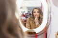 Young woman paints lips with lipstick in front of a mirror in a cosmetics store. Beautiful blonde in a beige coat. Fashion, style Royalty Free Stock Photo