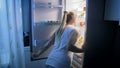 Young woman in pajams opens refrigerator at night and looking for food