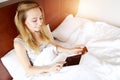 Young woman paying money for travel holds debit credit card and tablet laying in white bed at home with sunshine Royalty Free Stock Photo