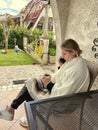 A young woman with a phone, covered with a blanket, sits on the terrace near the house and drinks tea.