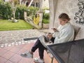 A young woman with a phone, covered with a blanket, sits on the terrace near the house and drinks tea.