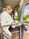 A young woman with a phone, covered with a blanket, sits on the terrace near the house and drinks tea.