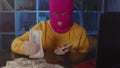 Young woman in pink balaclava collects money from table in office. Hacker in mask with dollar bills while sitting in