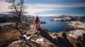 A young woman in a pink dress and hat sits on the edge of a cliff overlooking the fjord in Norway