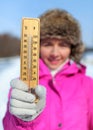 Young woman in pink jacket holds wooden thermometer outside on sunny but cold day, illustrating weather with freezing temperatures