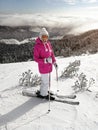 Young woman in pink jacket, skis, ski poles, goggles and hat, posing on the snow covered piste, sun shining to forest behind her Royalty Free Stock Photo