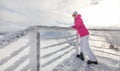 Young woman in pink ski jacket, boots gloves hat and goggles leaning on crystal snow covered fence rail, looking back. Sun shining Royalty Free Stock Photo