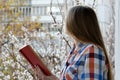 Young woman in plaid shirt with a book in hands stands by the window, spring city