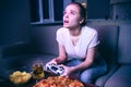 Young woman playing game at night. Emotional distracted girl look up. Holding gamepad in hands. Pizza with chips and