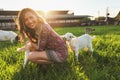 Young woman playing with goat kids on green spring meadow, smiling as animal chews her dress. Wide angle photo in strong sun Royalty Free Stock Photo