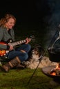 Young woman plays guitar in nature. Kettle is steaming over the campfire. Her tricolor Australian Shepherd watches and