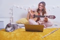 Young woman plays the guitar while sitting on the bed with a cat, online music lessons. Girl with a musical instrument in her