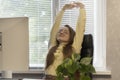 Young woman pleasantly smiling raising her hands upright while sitting at workplace. Secretary doing warm-up after work