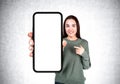 Young woman pointing at large phone display on grey concrete bac Royalty Free Stock Photo